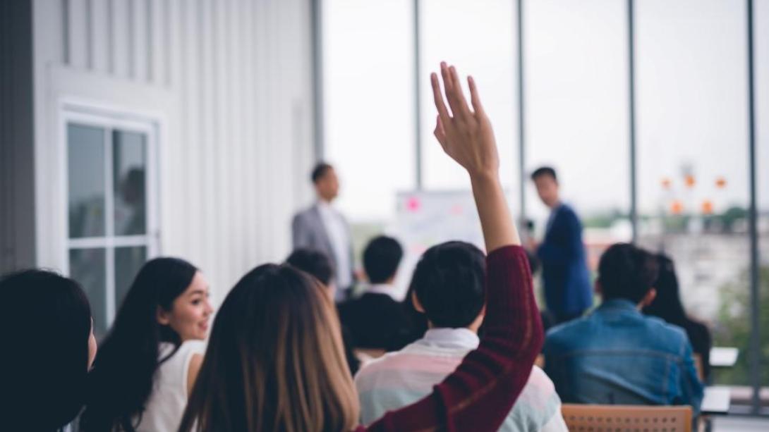Woman raising hand in presentation surrounded by colleagues in a room with large windows