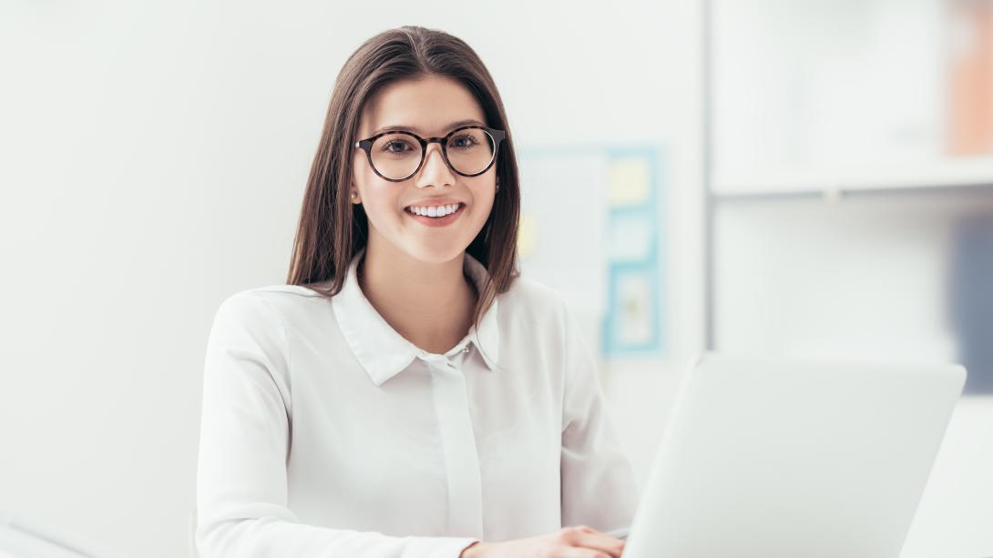 Young woman sitting at a desk typing on a laptop computer while looking at the camera and smiling