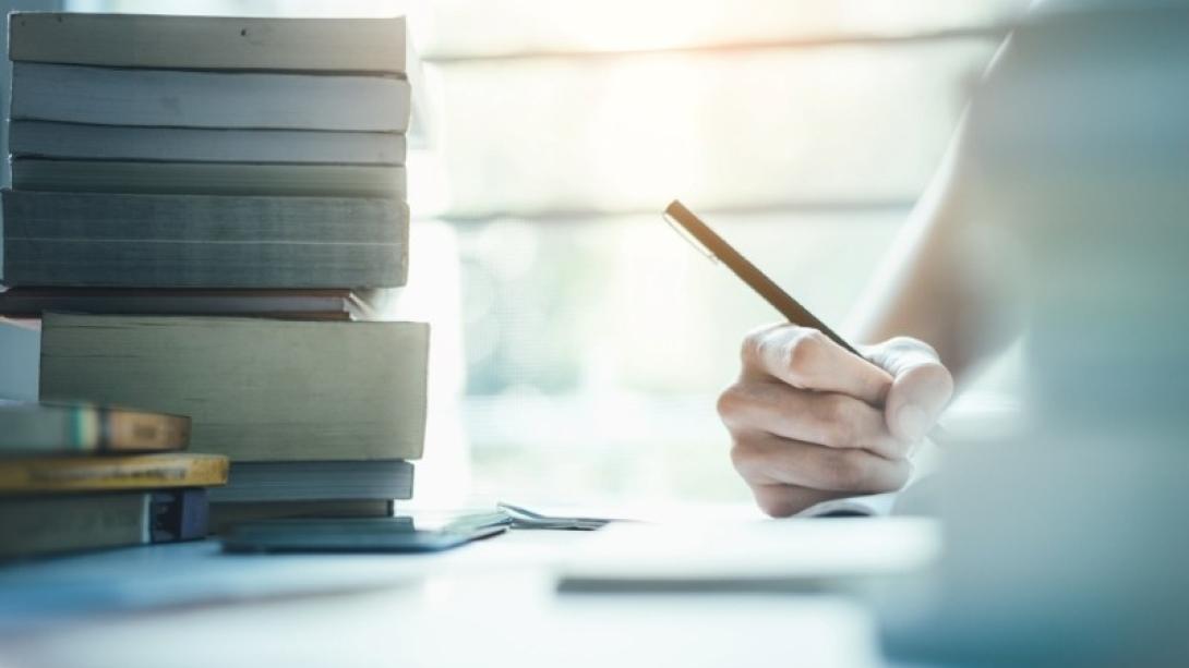 Person's hand holding a pen and writing in notepad on deck while surrounded by books