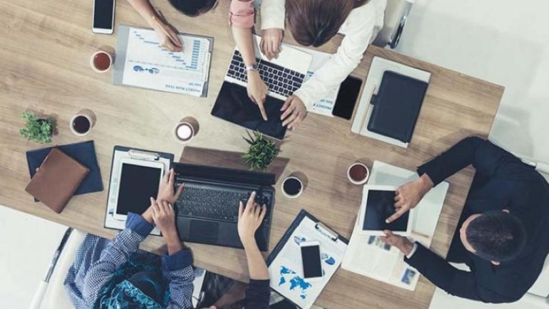 Birds eye view of five people working at a desk together on laptops and having a conversation