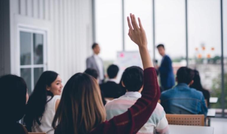 Woman raising hand in presentation surrounded by colleagues in a room with large windows