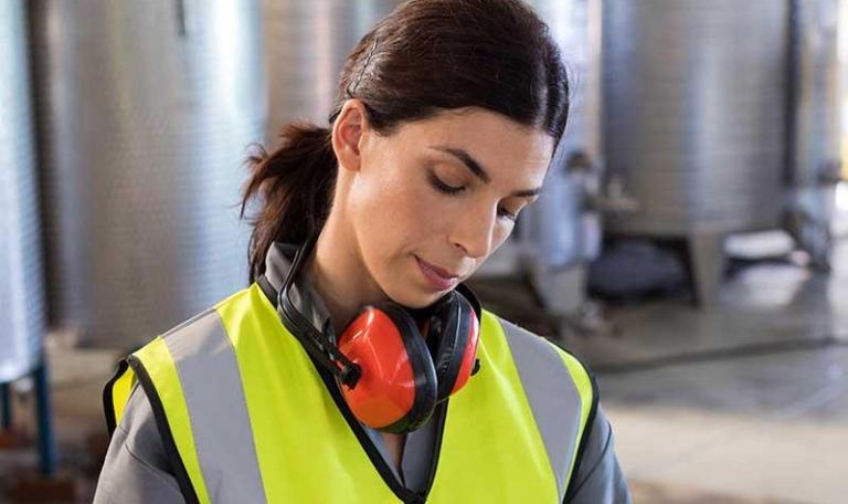 Female worker in warehouse looking at clipboard in a yellow high-vis vest with protective noise cancelling headphones