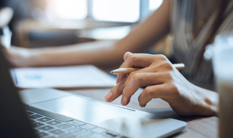 Shadowy image of hand of a woman poised over a laptop keyboard, pen between fingers.