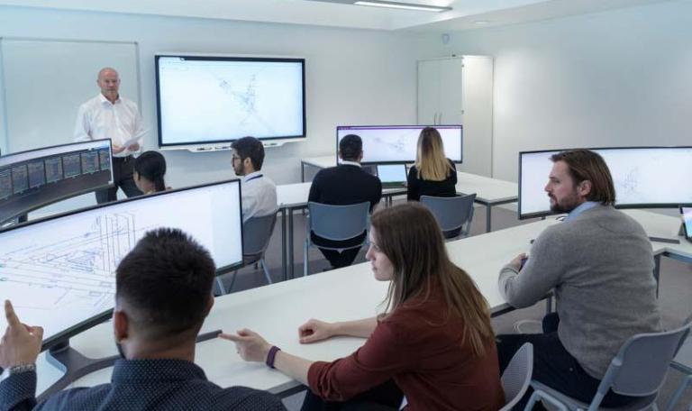 Classroom full of students sitting at desks with large screen computers with teacher presenting at front of room