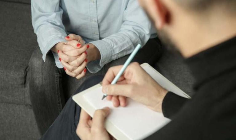 Counselling services offered to person that we cannot see their face and sitting on a couch, person sitting opposite writing in a notebook while listening