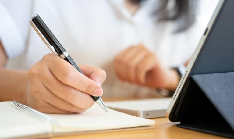 Person writing in notebook while looking at iPad filling out a form
