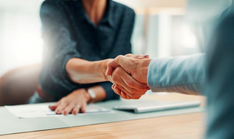 Two people shaking hands across a desk in an office space.