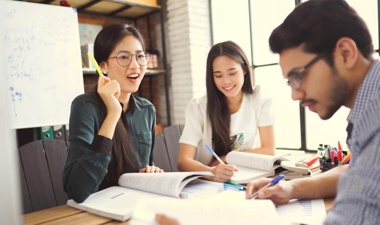 Three students sitting around a desk while reading books, studying and helping to prepare
