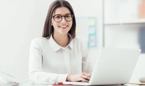Young woman sitting at a desk typing on a laptop computer while looking at the camera and smiling