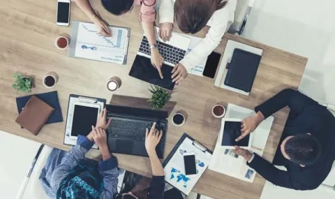 Birds eye view of five people working at a desk together on laptops and having a conversation
