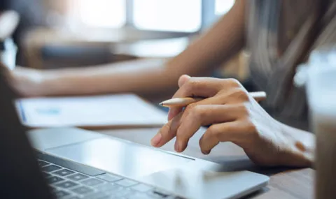 Shadowy image of hand of a woman poised over a laptop keyboard, pen between fingers.