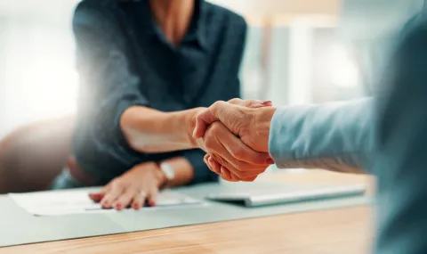 Two people shaking hands across a desk in an office space.
