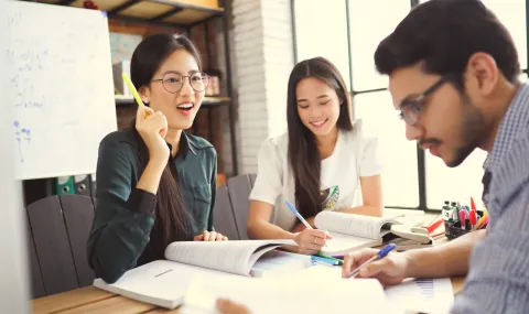 Three students sitting around a desk while reading books, studying and helping to prepare